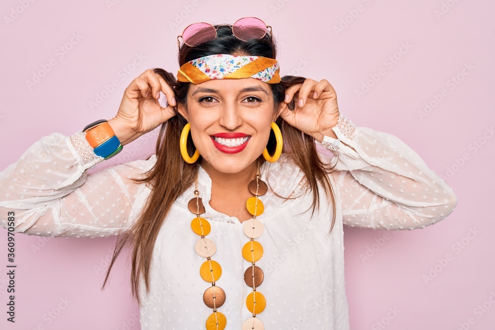 Young hispanic hippie woman wearing fashion boho style and sunglasses over pink background Smiling pulling ears with fingers, funny gesture. Audition problem Young hispanic hippie woman wearing fashion boho style and sunglasses over pink background Smiling pulling ears with fingers, funny gesture. Audition problem