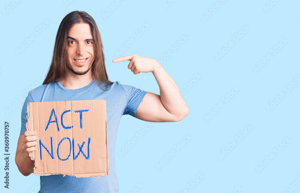 Young adult man with long hair holding act now banner pointing finger to one self smiling happy and proud Young adult man with long hair holding act now banner pointing finger to one self smiling happy and proud