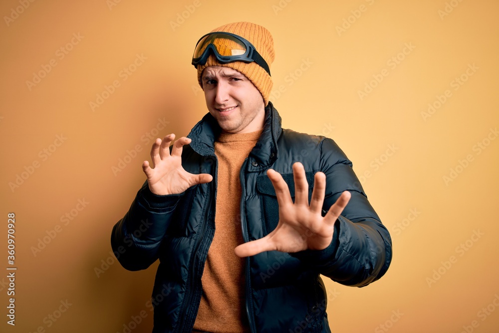Young handsome caucasian man wearing hat, coat and ski glasses for winter and snow weather disgusted expression, displeased and fearful doing disgust face because aversion reaction. With hands raised Young handsome caucasian man wearing hat, coat and ski glasses for winter and snow weather disgusted expression, displeased and fearful doing disgust face because aversion reaction. With hands raised