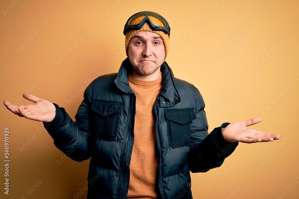 Young handsome caucasian man wearing hat, coat and ski glasses for winter and snow weather clueless and confused expression with arms and hands raised. Doubt concept. Young handsome caucasian man wearing hat, coat and ski glasses for winter and snow weather clueless and confused expression with arms and hands raised. Doubt concept.