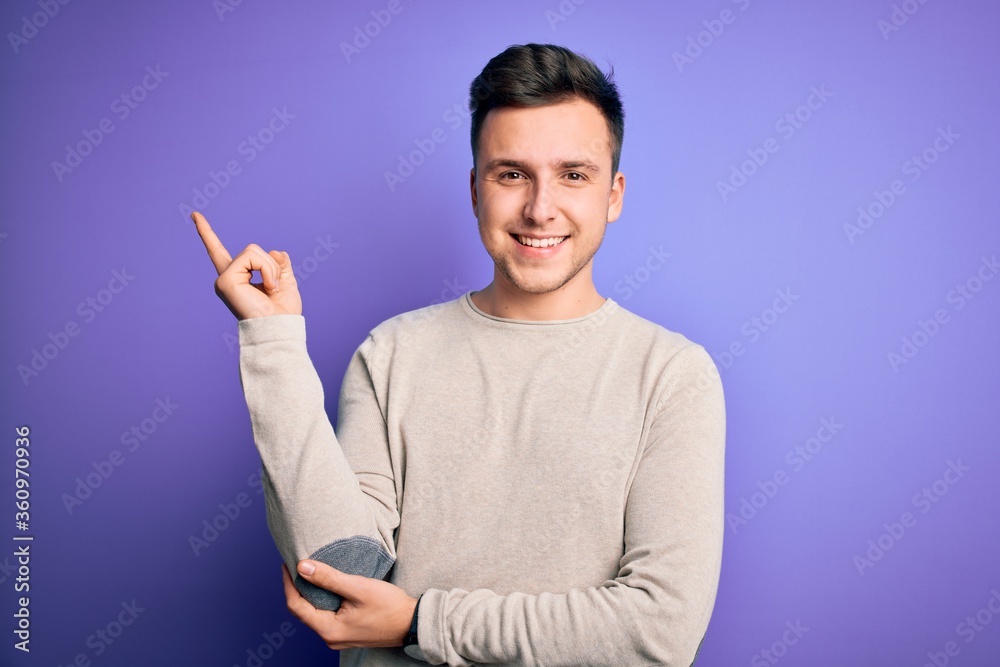 Young handsome caucasian man wearing casual sweater over purple isolated background with a big smile on face, pointing with hand and finger to the side looking at the camera. Young handsome caucasian man wearing casual sweater over purple isolated background with a big smile on face, pointing with hand and finger to the side looking at the camera.