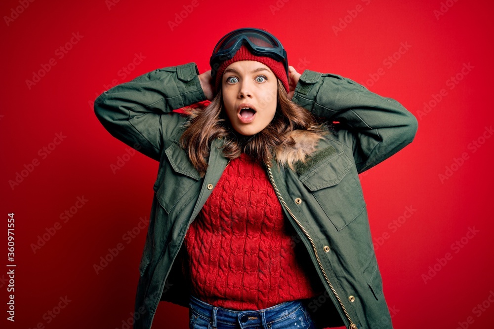 Young blonde girl wearing ski glasses and winter coat for ski weather over red background Crazy and scared with hands on head, afraid and surprised of shock with open mouth