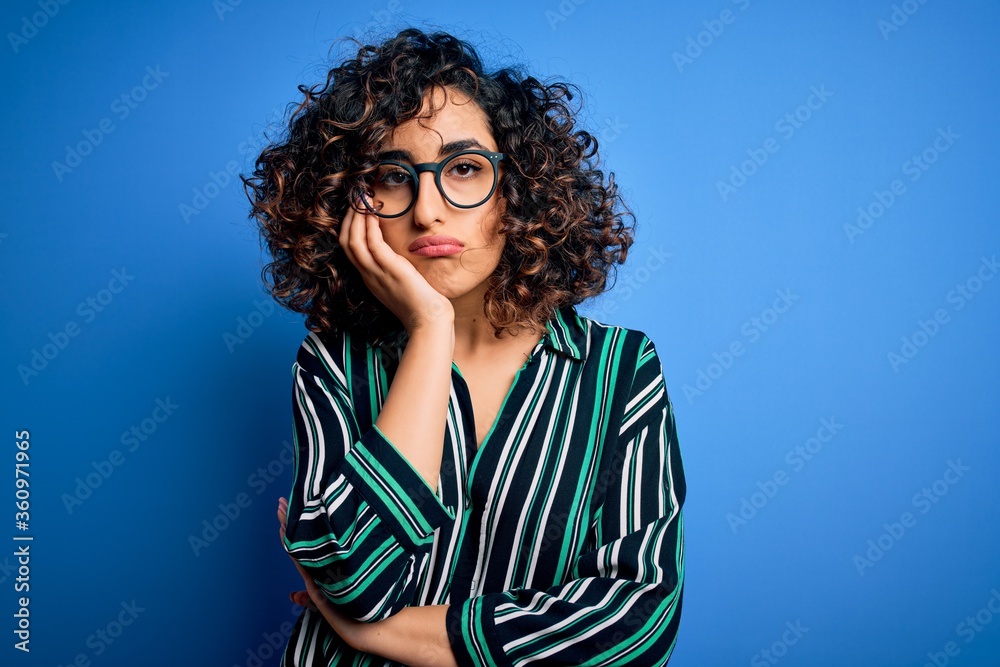 Young beautiful curly arab woman wearing striped shirt and glasses over blue background thinking looking tired and bored with depression problems with crossed arms. Young beautiful curly arab woman wearing striped shirt and glasses over blue background thinking looking tired and bored with depression problems with crossed arms.