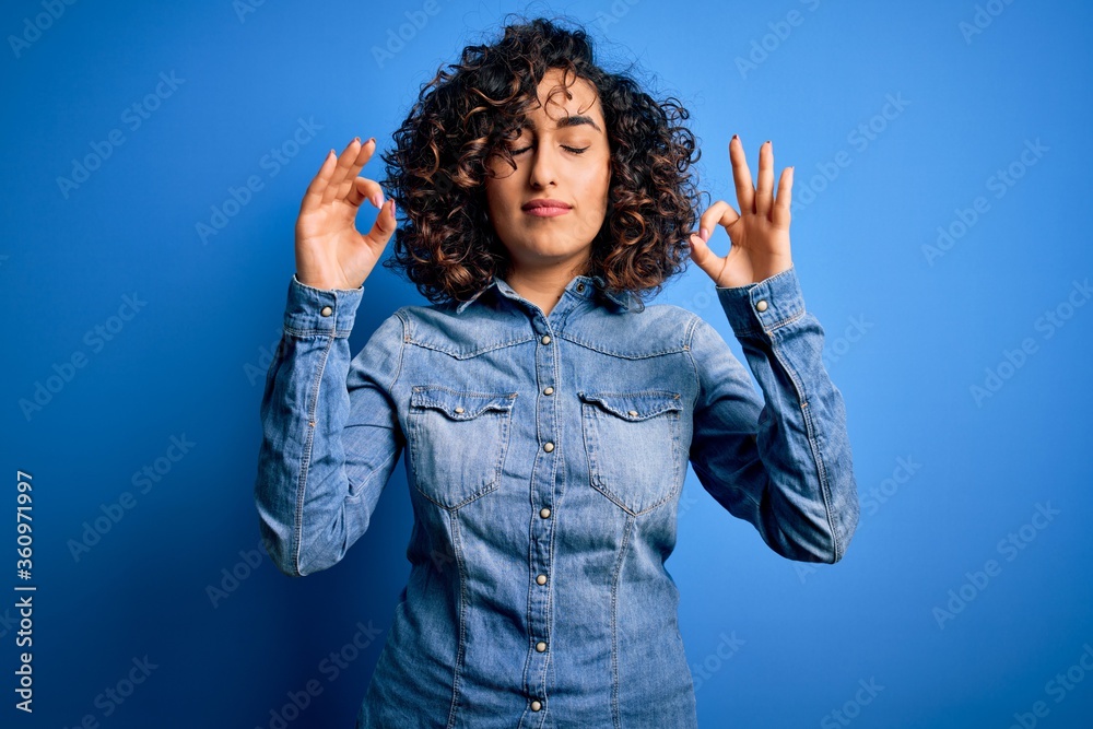 Young beautiful curly arab woman wearing casual denim shirt standing over blue background relax and smiling with eyes closed doing meditation gesture with fingers. Yoga concept. Young beautiful curly arab woman wearing casual denim shirt standing over blue background relax and smiling with eyes closed doing meditation gesture with fingers. Yoga concept.