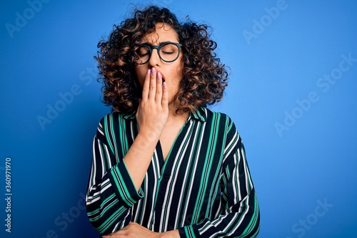 Young beautiful curly arab woman wearing striped shirt and glasses over blue background bored yawning tired covering mouth with hand. Restless and sleepiness.