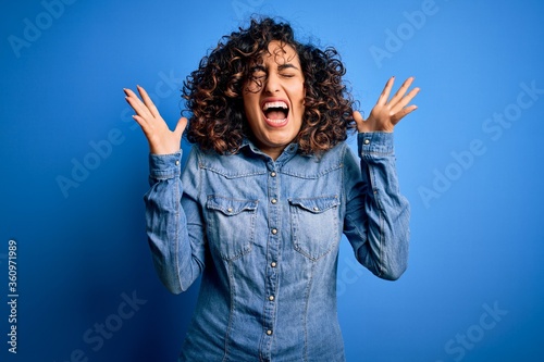 Young beautiful curly arab woman wearing casual denim shirt standing over blue background celebrating mad and crazy for success with arms raised and closed eyes screaming excited. Winner concept