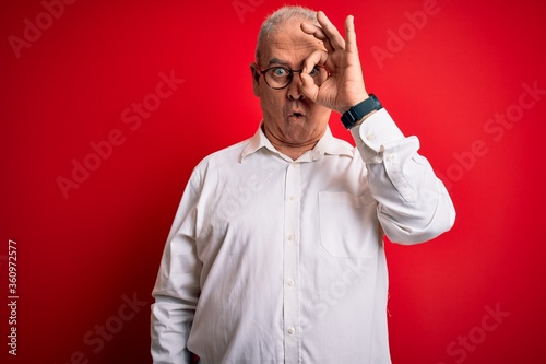 Middle age handsome hoary man wearing casual shirt and glasses over red background doing ok gesture shocked with surprised face, eye looking through fingers. Unbelieving expression.