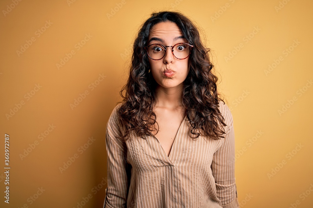 Beautiful woman with curly hair wearing striped shirt and glasses over yellow background puffing cheeks with funny face. Mouth inflated with air, crazy expression.