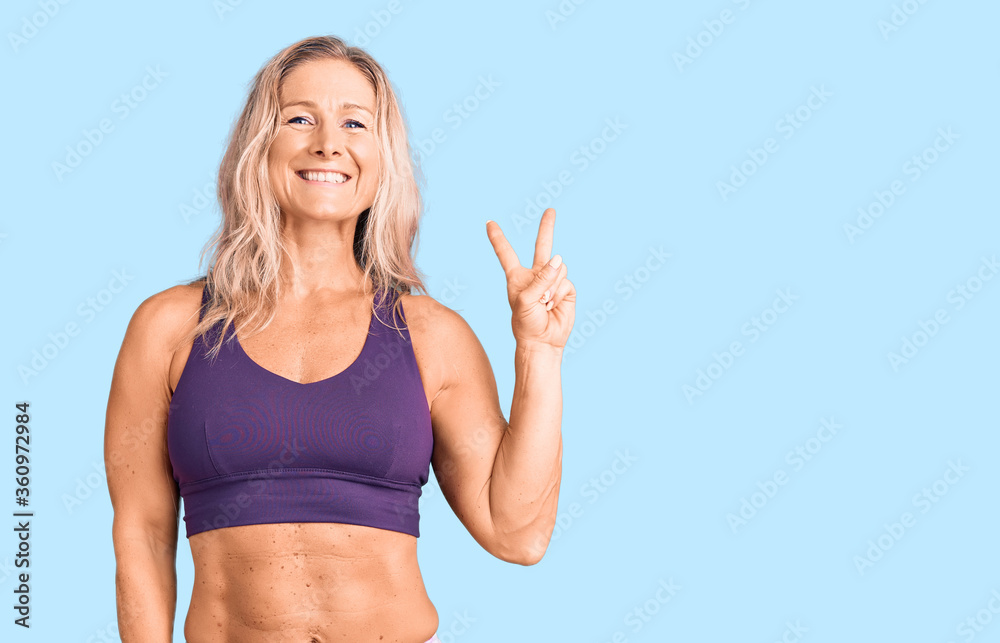 Middle age fit blonde woman wearing sportswear smiling with happy face winking at the camera doing victory sign. number two. Middle age fit blonde woman wearing sportswear smiling with happy face winking at the camera doing victory sign. number two.