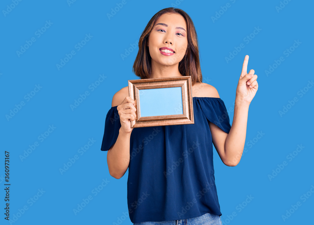 Young beautiful chinese girl holding empty frame surprised with an idea or question pointing finger with happy face, number one Young beautiful chinese girl holding empty frame surprised with an idea or question pointing finger with happy face, number one