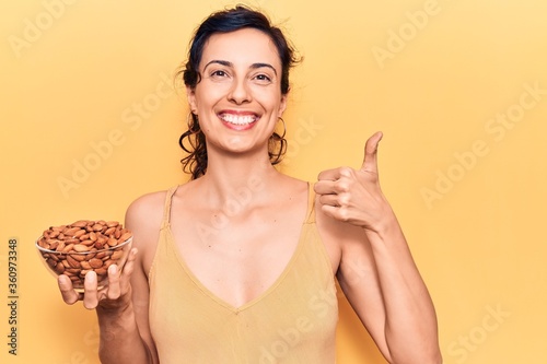 Young beautiful hispanic woman holding bowl with almonds smiling happy and positive, thumb up doing excellent and approval sign