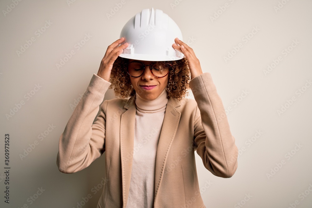 Young african american curly architect woman wearing safety helmet and glasses suffering from headache desperate and stressed because pain and migraine. Hands on head. Young african american curly architect woman wearing safety helmet and glasses suffering from headache desperate and stressed because pain and migraine. Hands on head.