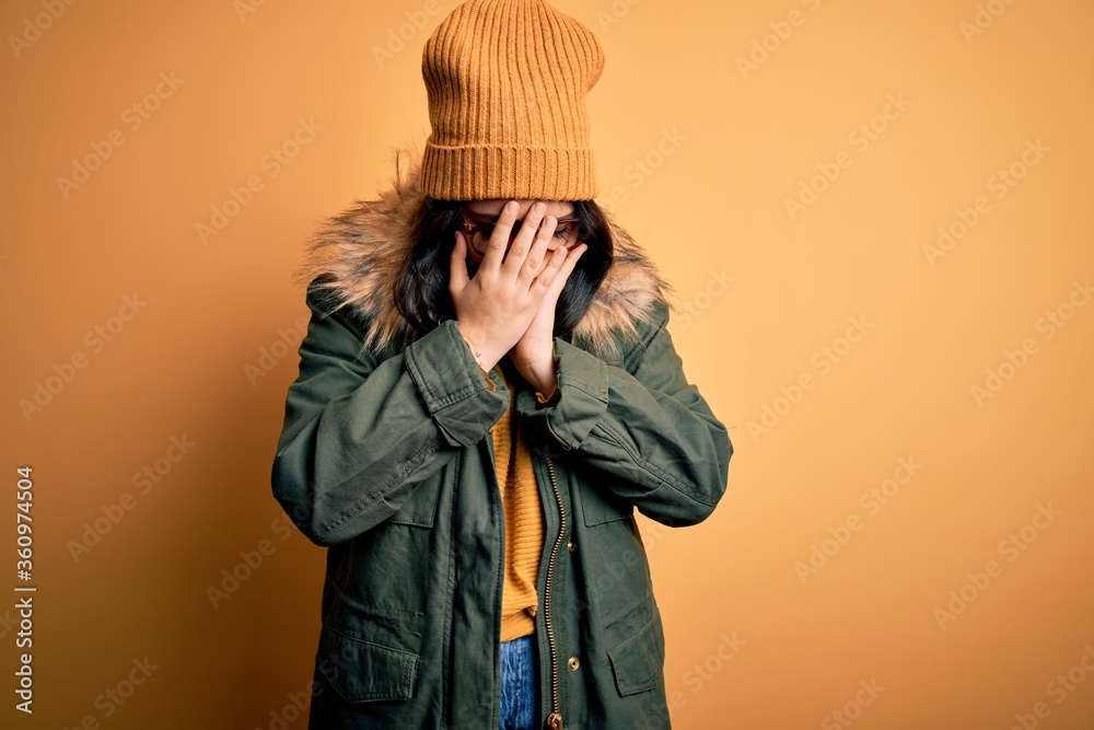 Young brunette woman wearing glasses and winter coat with hat over yellow isolated background with sad expression covering face with hands while crying. Depression concept. Young brunette woman wearing glasses and winter coat with hat over yellow isolated background with sad expression covering face with hands while crying. Depression concept.