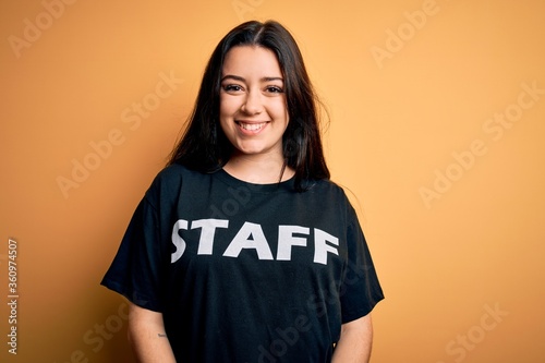 Young brunette worker woman wearing staff t-shirt as uniform over yellow isolated background with a happy face standing and smiling with a confident smile showing teeth