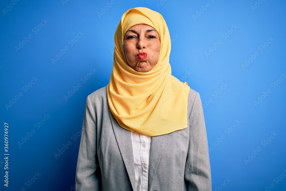 Middle age brunette business woman wearing muslim traditional hijab over blue background puffing cheeks with funny face. Mouth inflated with air, crazy expression.