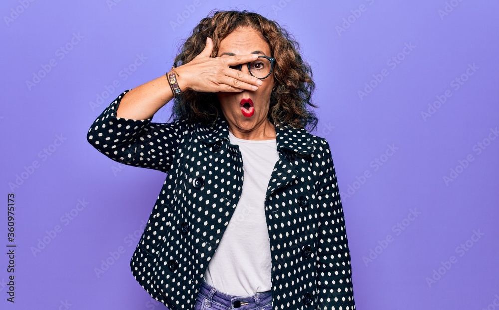 Middle age beautiful woman wearing casual coat and glasses over isolated purple background peeking in shock covering face and eyes with hand, looking through fingers afraid Middle age beautiful woman wearing casual coat and glasses over isolated purple background peeking in shock covering face and eyes with hand, looking through fingers afraid