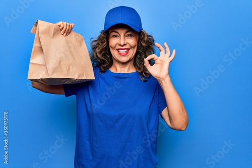 Middle age beautiful delivery woman holding deliver paper bag with takeaway food doing ok sign with fingers, smiling friendly gesturing excellent symbol