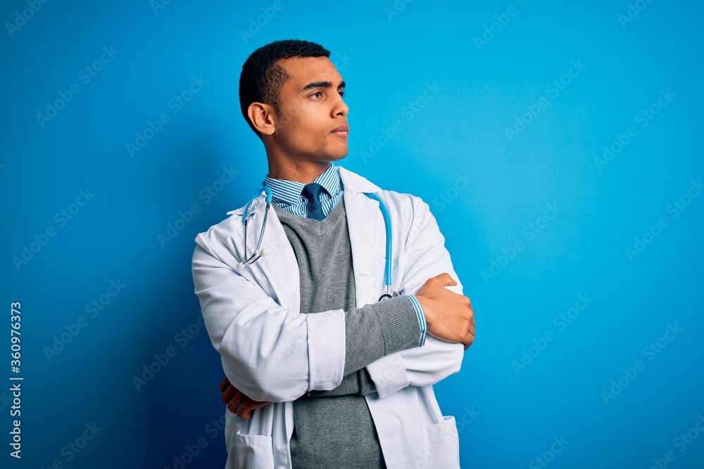 Handsome african american doctor man wearing coat and stethoscope over blue background looking to the side with arms crossed convinced and confident Handsome african american doctor man wearing coat and stethoscope over blue background looking to the side with arms crossed convinced and confident