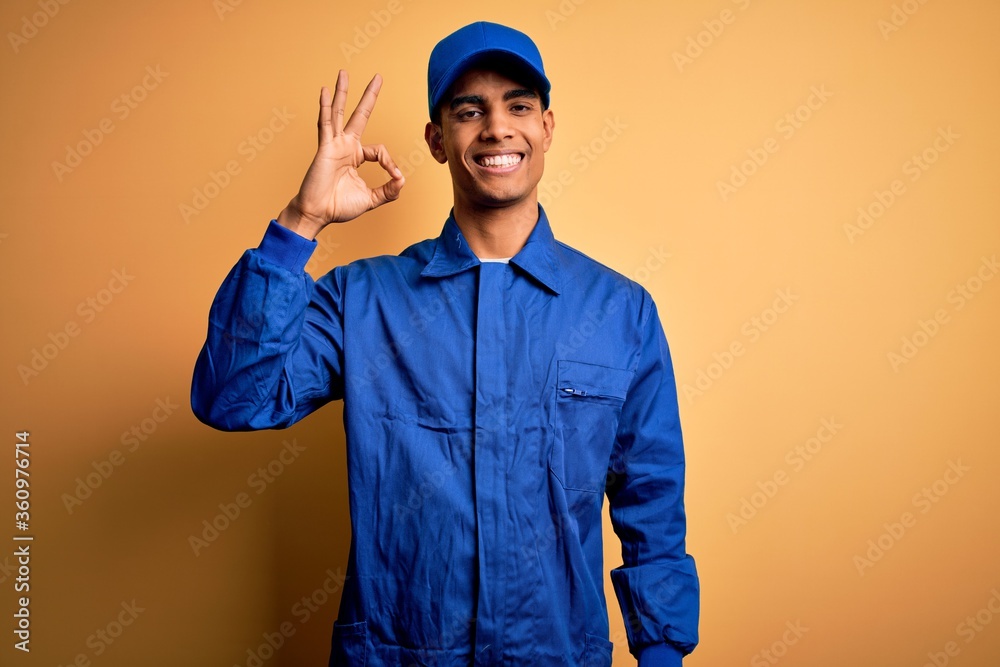 Young african american mechanic man wearing blue uniform and cap over yellow background smiling positive doing ok sign with hand and fingers. Successful expression. Young african american mechanic man wearing blue uniform and cap over yellow background smiling positive doing ok sign with hand and fingers. Successful expression.