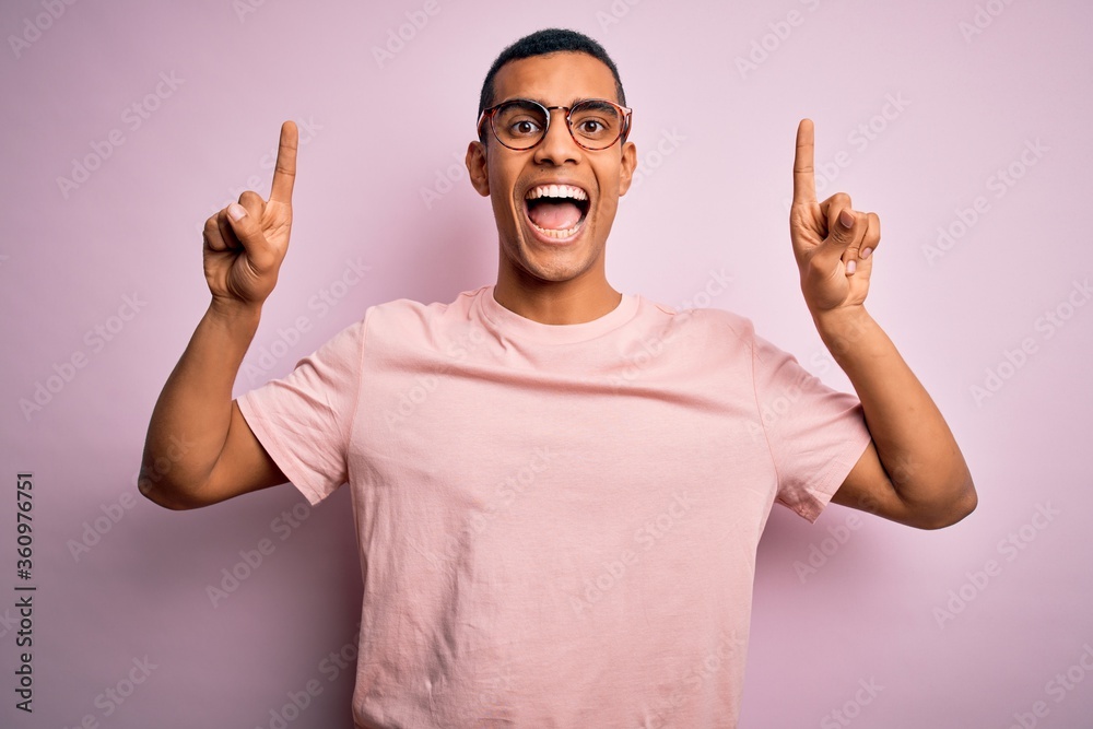 Handsome african american man wearing casual t-shirt and glasses over pink background smiling amazed and surprised and pointing up with fingers and raised arms. Handsome african american man wearing casual t-shirt and glasses over pink background smiling amazed and surprised and pointing up with fingers and raised arms.
