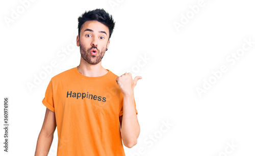 Young hispanic man wearing t shirt with happiness word message surprised pointing with hand finger to the side, open mouth amazed expression.