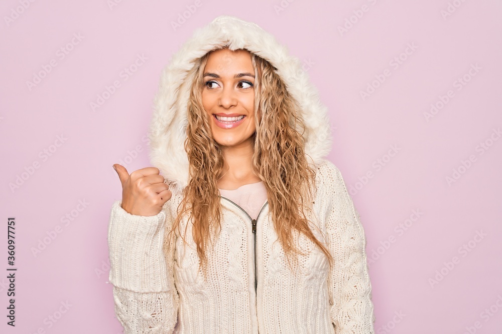 Young beautiful blonde woman wearing casual sweater with hood over isolated pink background smiling with happy face looking and pointing to the side with thumb up. Young beautiful blonde woman wearing casual sweater with hood over isolated pink background smiling with happy face looking and pointing to the side with thumb up.