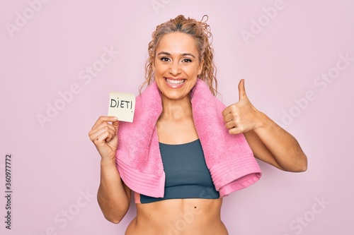 Beautiful blonde sporty woman doing sport wearing towel holding reminder with diet message Smiling happy and positive, thumb up doing excellent and approval sign