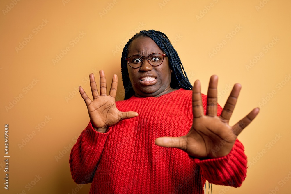 African american plus size woman with braids wearing casual sweater over yellow background afraid and terrified with fear expression stop gesture with hands, shouting in shock. Panic concept. African american plus size woman with braids wearing casual sweater over yellow background afraid and terrified with fear expression stop gesture with hands, shouting in shock. Panic concept.
