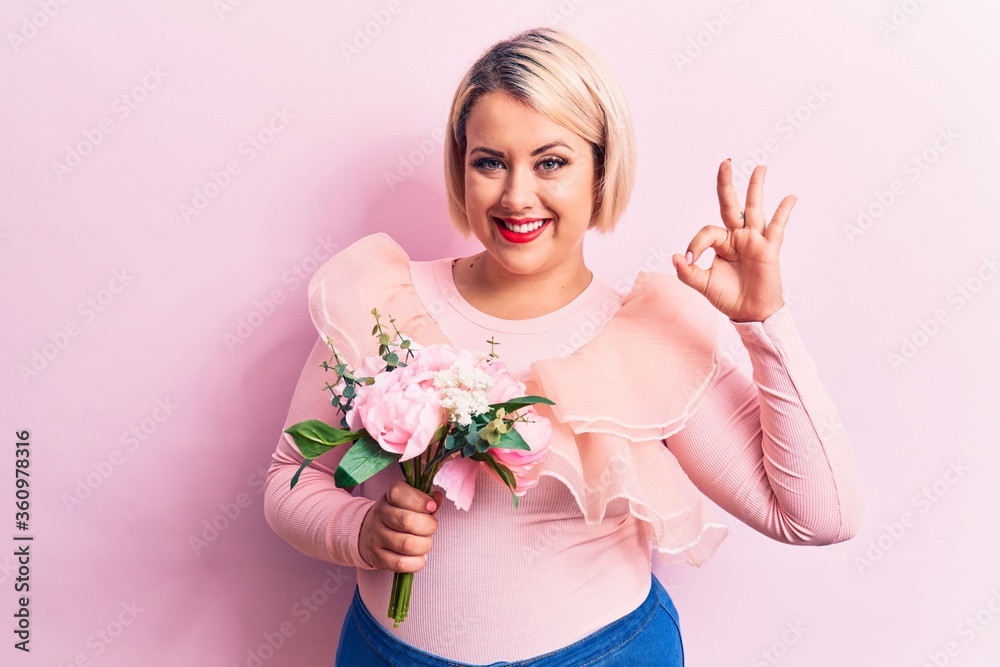 Beautiful blonde plus size woman holding bouquet of pink flowers over isolated background doing ok sign with fingers, smiling friendly gesturing excellent symbol