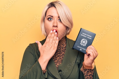 Young beautiful plus size tourist woman holding Italy Itaian passport over yellow background covering mouth with hand, shocked and afraid for mistake. Surprised expression photo