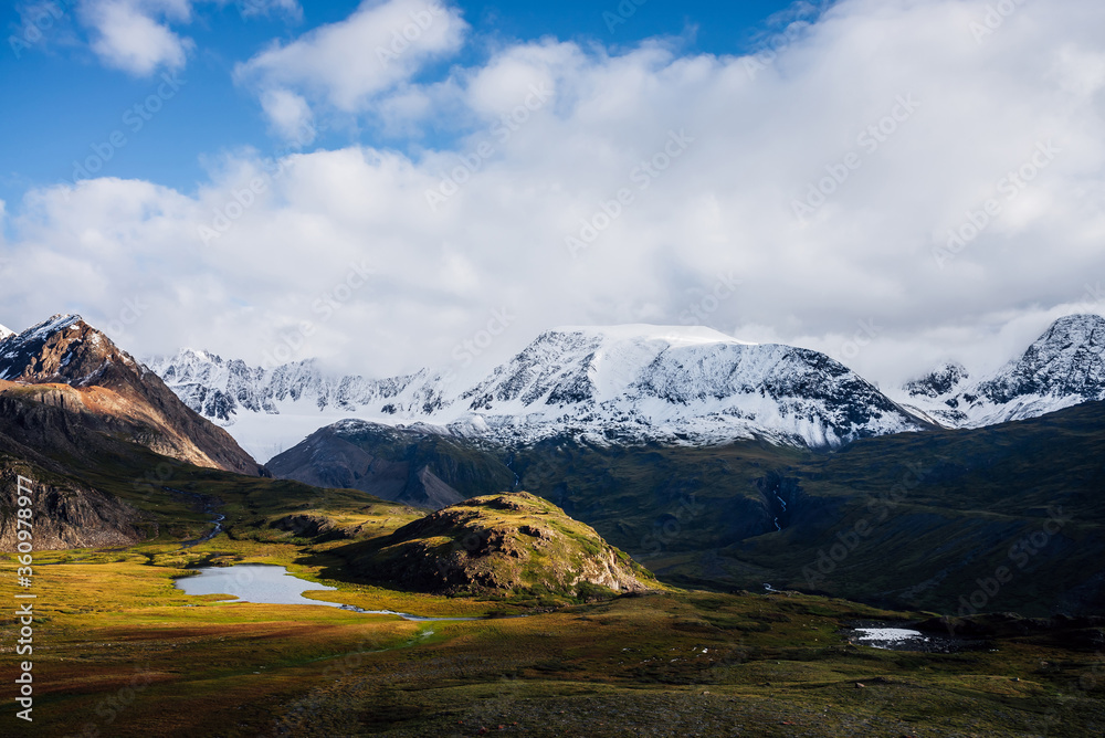 Awesome picturesque view to mountain range in sunny day. Beautiful green highland relief with glacier, lake, river, white snowy tops and low clouds in sunlight. Scenic landscape with snowy mount ridge