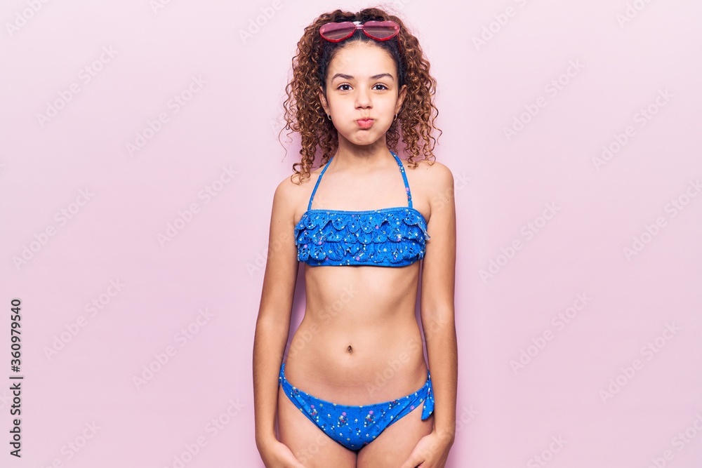 Naklejka premium Beautiful kid girl with curly hair wearing bikini and sunglasses puffing cheeks with funny face. mouth inflated with air, crazy expression.
