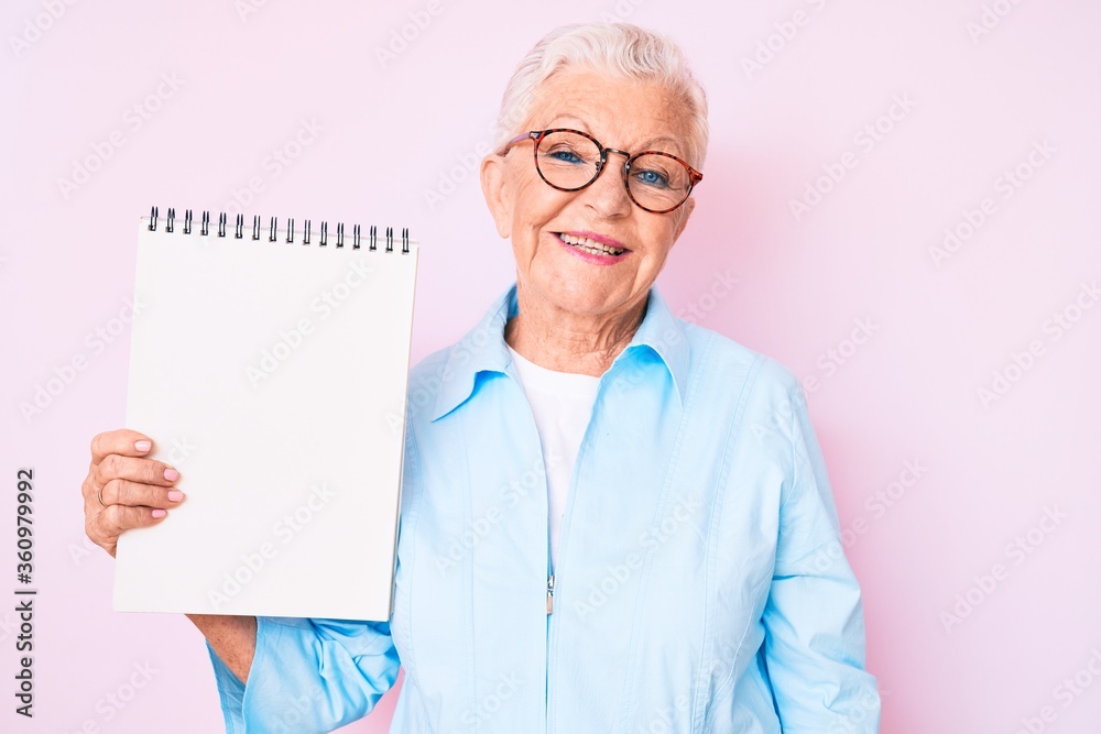 Senior beautiful woman with blue eyes and grey hair holding notebook looking positive and happy standing and smiling with a confident smile showing teeth Senior beautiful woman with blue eyes and grey hair holding notebook looking positive and happy standing and smiling with a confident smile showing teeth