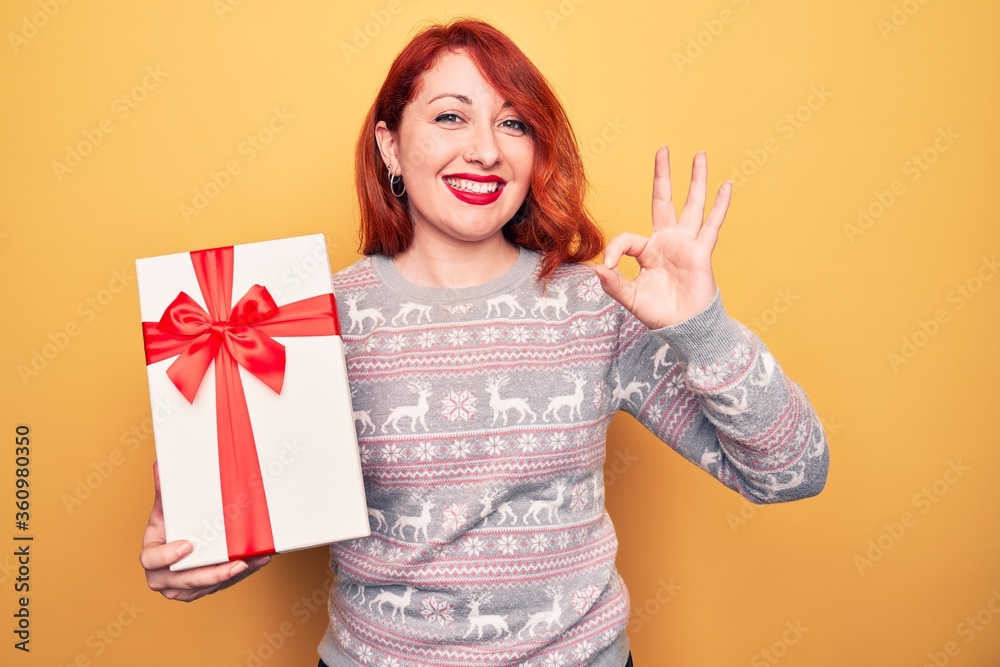 Young beautiful redhead woman holding christmas gift over isolated yellow background doing ok sign with fingers, smiling friendly gesturing excellent symbol Young beautiful redhead woman holding christmas gift over isolated yellow background doing ok sign with fingers, smiling friendly gesturing excellent symbol