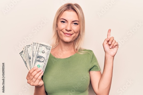 Young beautiful blonde woman holding bunch of dollars banknotes over white background smiling with an idea or question pointing finger with happy face, number one