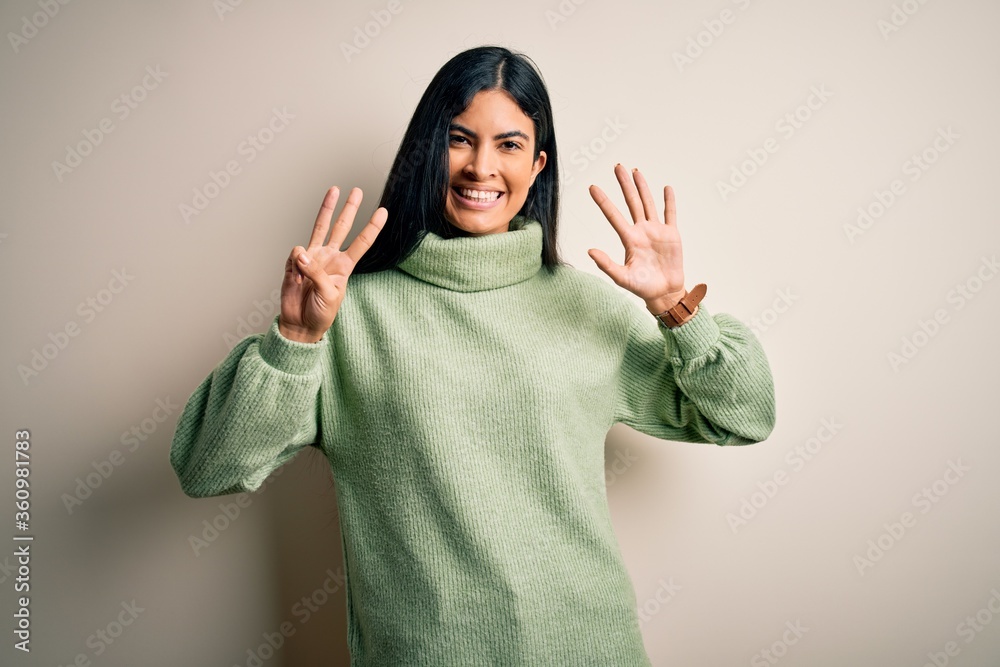 Young beautiful hispanic woman wearing green winter sweater over isolated background showing and pointing up with fingers number eight while smiling confident and happy. Young beautiful hispanic woman wearing green winter sweater over isolated background showing and pointing up with fingers number eight while smiling confident and happy.