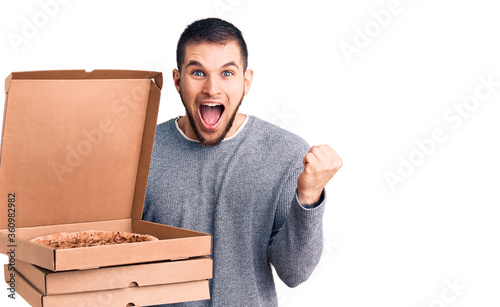 Young handsome man holding delivery italian pizza boxes screaming proud, celebrating victory and success very excited with raised arms photo