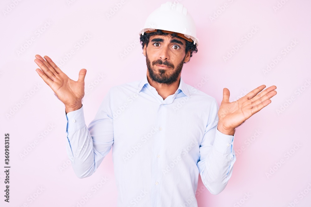 Handsome young man with curly hair and bear wearing architect hardhat clueless and confused with open arms, no idea and doubtful face. Handsome young man with curly hair and bear wearing architect hardhat clueless and confused with open arms, no idea and doubtful face.