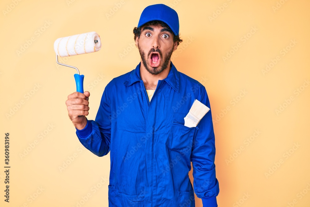Handsome young man with curly hair and bear wearing builder jumpsuit uniform holding paint roller scared and amazed with open mouth for surprise, disbelief face
