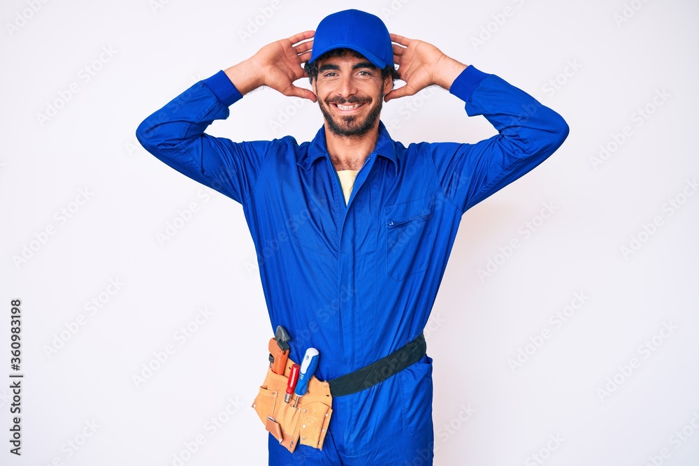 Handsome young man with curly hair and bear weaing handyman uniform relaxing and stretching, arms and hands behind head and neck smiling happy