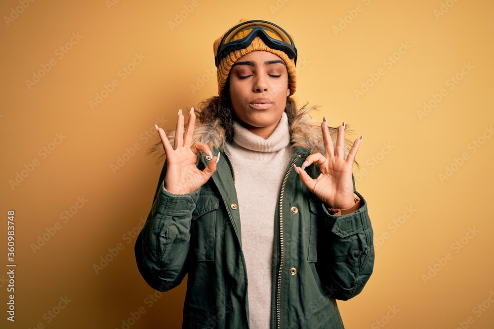 Young african american afro skier girl wearing snow sportswear and ski goggles relaxed and smiling with eyes closed doing meditation gesture with fingers. Yoga concept.