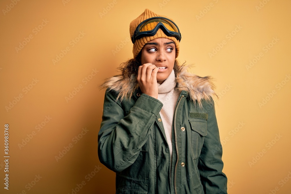 Young african american afro skier girl wearing snow sportswear and ski goggles looking stressed and nervous with hands on mouth biting nails. Anxiety problem. Young african american afro skier girl wearing snow sportswear and ski goggles looking stressed and nervous with hands on mouth biting nails. Anxiety problem.