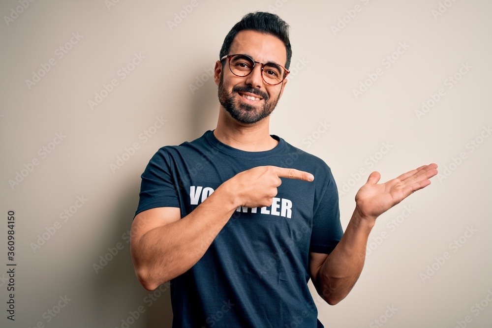 Handsome man with beard wearing t-shirt with volunteer message over white background amazed and smiling to the camera while presenting with hand and pointing with finger. Handsome man with beard wearing t-shirt with volunteer message over white background amazed and smiling to the camera while presenting with hand and pointing with finger.