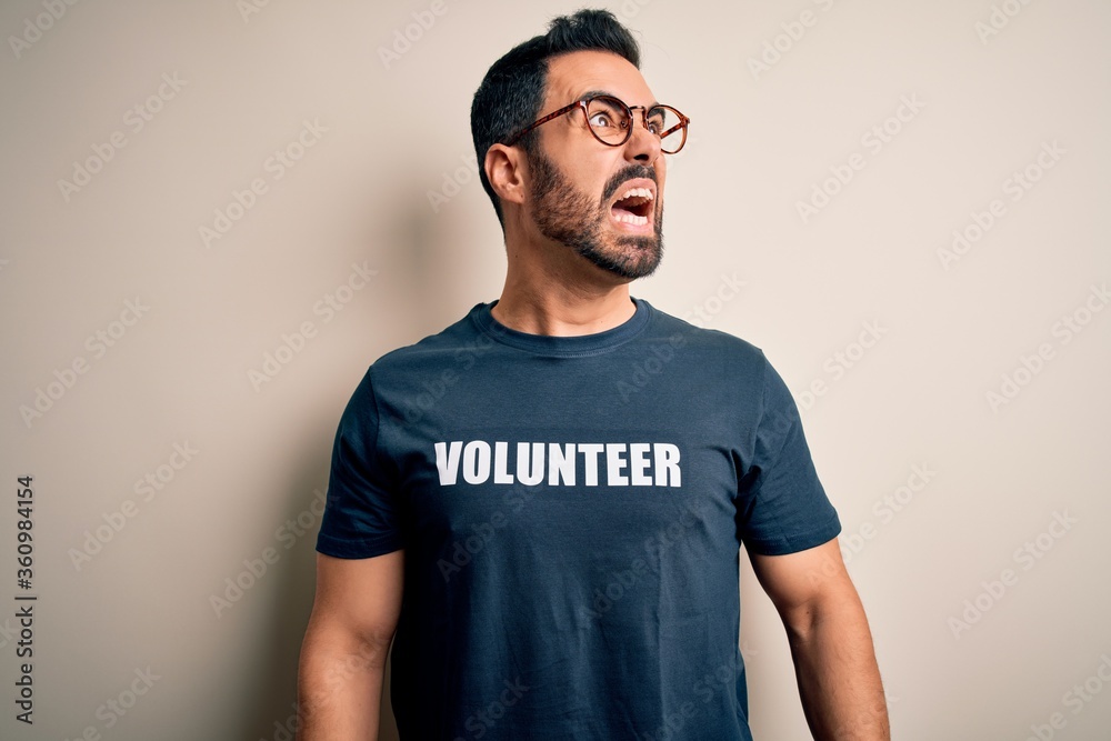 Handsome man with beard wearing t-shirt with volunteer message over white background angry and mad screaming frustrated and furious, shouting with anger. Rage and aggressive concept. Handsome man with beard wearing t-shirt with volunteer message over white background angry and mad screaming frustrated and furious, shouting with anger. Rage and aggressive concept.
