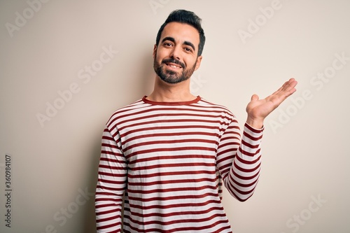 Young handsome man with beard wearing casual striped t-shirt standing over white background smiling cheerful presenting and pointing with palm of hand looking at the camera.