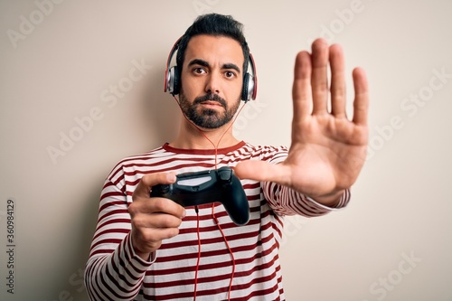 Young handsome gamer man with beard playing video game using joystick and headphones with open hand doing stop sign with serious and confident expression, defense gesture
