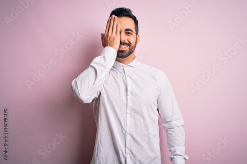 Young handsome man with beard wearing casual shirt standing over pink background covering one eye with hand, confident smile on face and surprise emotion.
