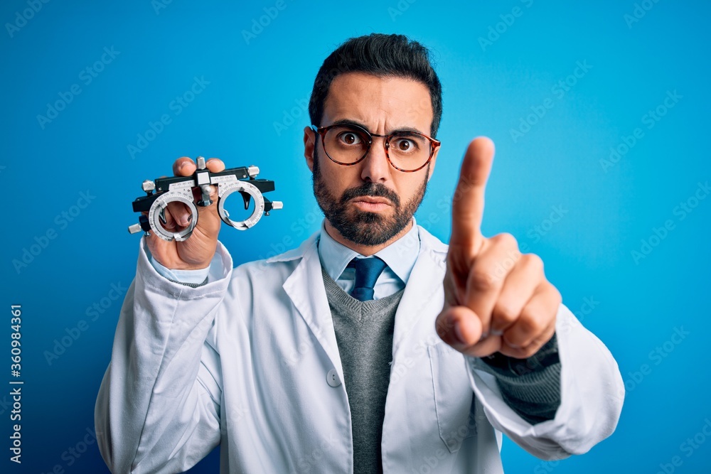 Young handsome optical man with beard holding optometry glasses over blue background Pointing with finger up and angry expression, showing no gesture Young handsome optical man with beard holding optometry glasses over blue background Pointing with finger up and angry expression, showing no gesture