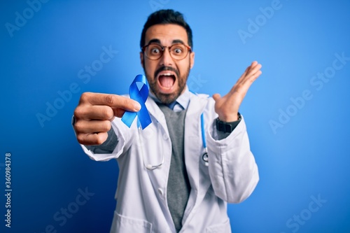 Young handsome doctor man with beard wearing stethoscope holding blue cancer ribbon very happy and excited, winner expression celebrating victory screaming with big smile and raised hands