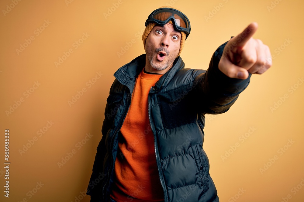 Young handsome skier man skiing wearing snow sportswear using ski goggles Pointing with finger surprised ahead, open mouth amazed expression, something on the front
